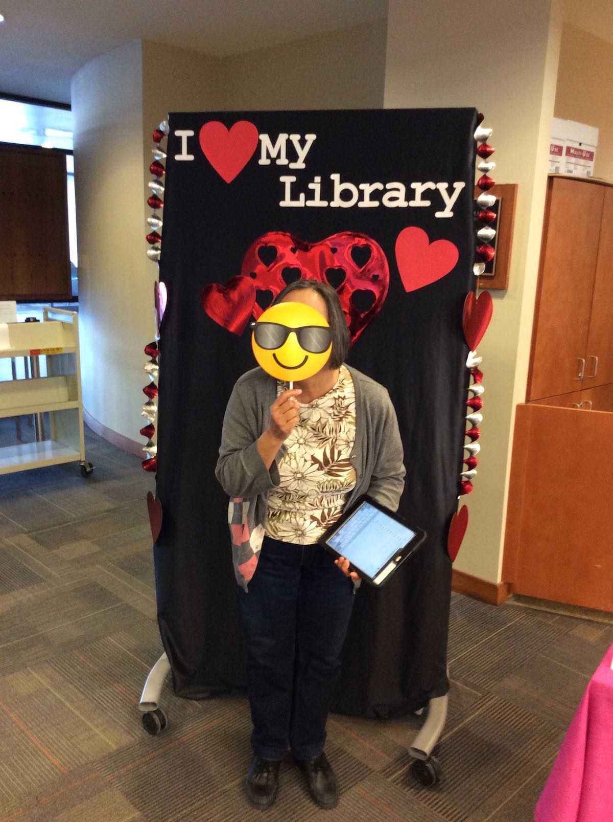 Library User Services Assistant Cecilia Gallardo stands in front of the I love my library backdrop holding up a prop of a smiling sunglasses emoji, covering her face with it